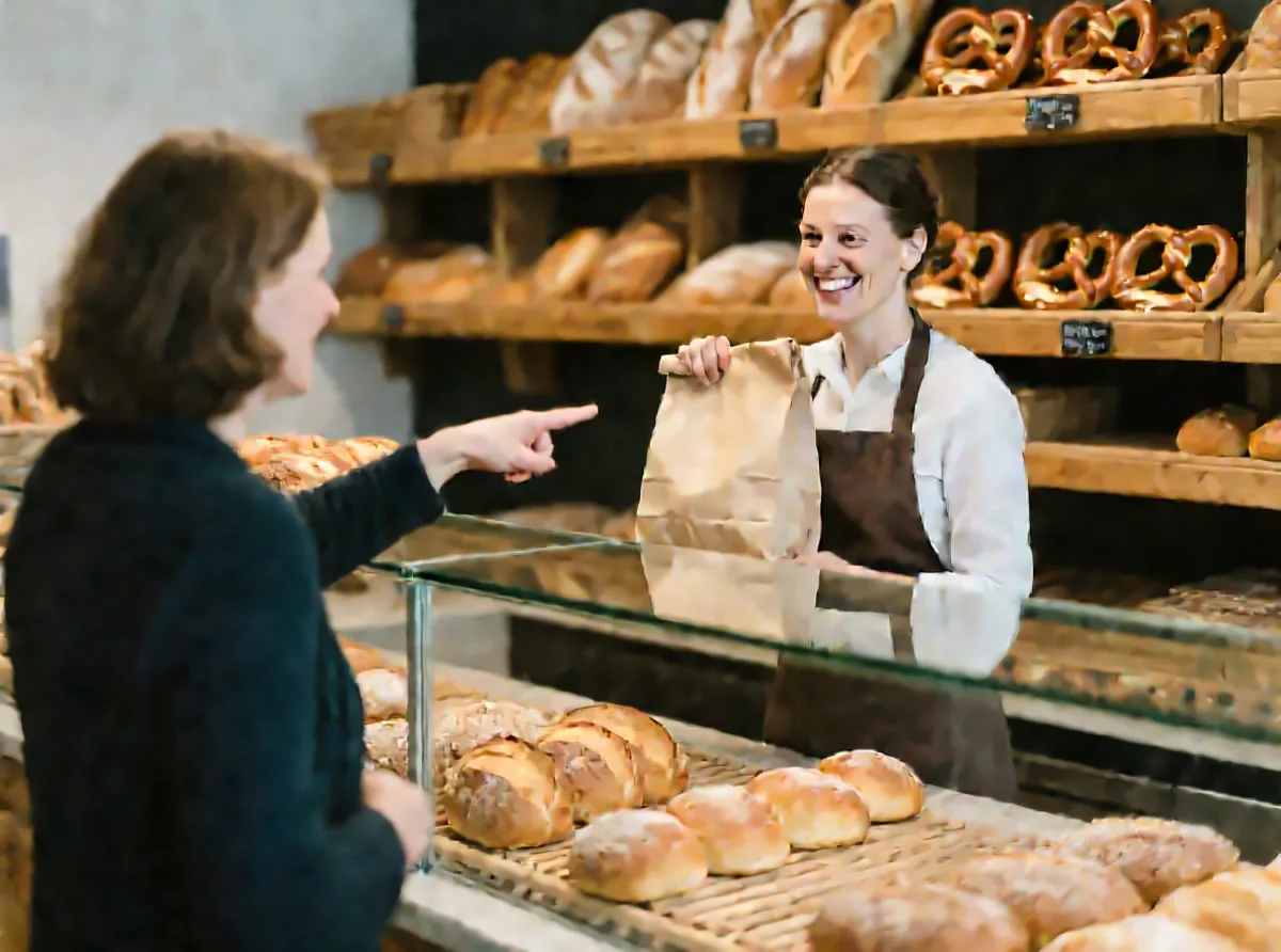 Bild 1: In einer Bäckerei zeigt eine Kundin auf Brötchen, eine Verkäuferin hält eine Papiertüte.